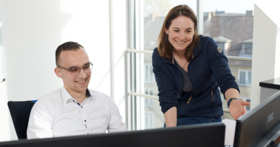 Kollegen, die am Arbeitsplatz lächelnd auf einen PC deuten. 