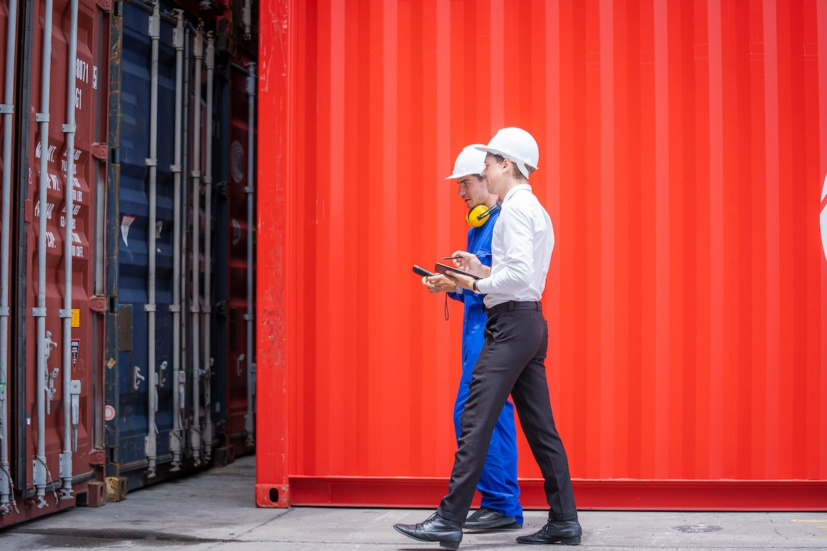 Zwei Männer mit Schutzhelmen gehen nebeneinander an großen Containern vorbei, einer trägt blaue Arbeitskleidung und der andere ein weißes Hemd mit schwarzer Hose